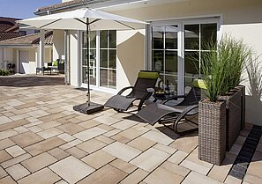 Zwei Liegestühle unter dem weißen Terrassenschirm laden ein die Sonnenstunden auf der mediterran gestalteten Terrasse zu genießen. Die Platten mit Farbspiel sind wild verlegt.
