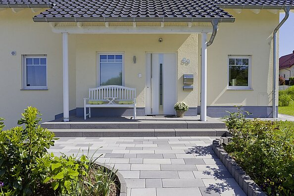 Der Eingangsbereich des klassischen Neubauhauses in zartem Gelb mit Vordach gestützt mit Säulen ist gestaltet mit zwei Treppenstufen und Pflaster mit Farbspiel verschiedener Grautöne. Auf dem Podest vor der Haustür passt noch eine weiße Holzbank hin!